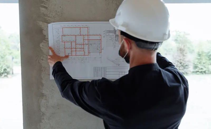 Person in a hard hat, seen from behind, reviewing a floor plan pinned to a concrete column